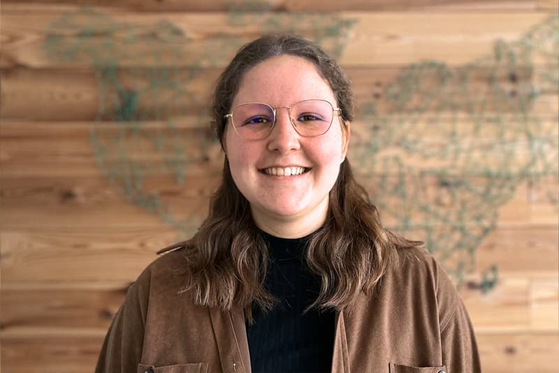 Eine lächelnde Person mit Brille und gewelltem Haar steht vor einer Holzwand mit einem Weltkartenmotiv.