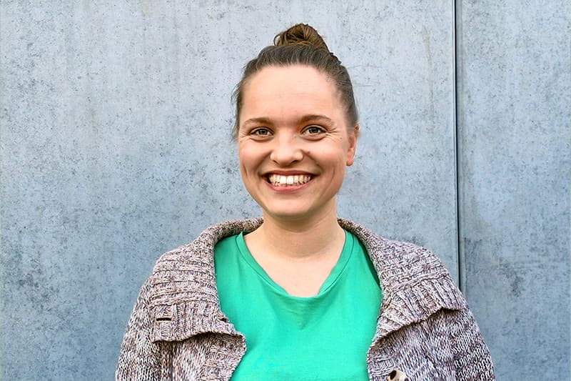 Eine Frau mit Dutt lächelt in die Kamera. Sie trägt ein grünes Hemd und einen Strickpullover und steht vor einer grauen Wand.