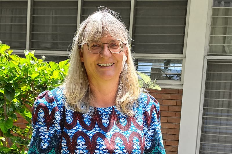 Eine lächelnde Frau mit langen blonden Haaren und Brille steht in einem blau gemusterten Oberteil draußen vor Fenstern und Grünanlagen.