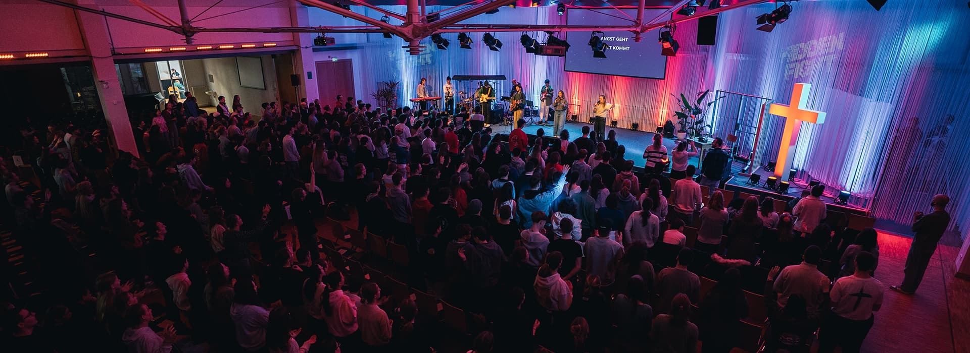 Eine große Menschenmenge nimmt an einem Gottesdienst mit einer Band auf der Bühne und einem leuchtenden Kreuz unter farbiger Beleuchtung teil.