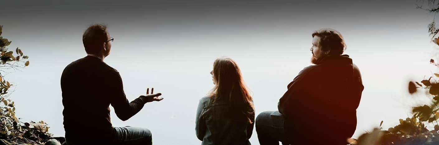 Drei Personen sitzen im Freien vor einem hellen, nebligen Hintergrund, unterhalten sich und sind von Laub umgeben.