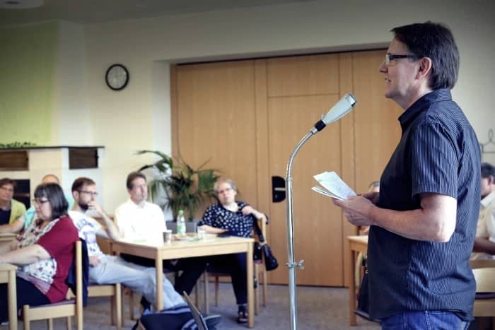 Ein Mann spricht in ein Mikrofon, während die an Tischen sitzenden Personen in einem Besprechungsraum aufmerksam zuhören.