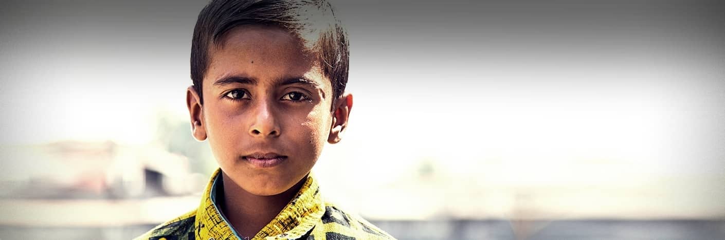 Ein Junge mit kurzen dunklen Haaren und einem gelb gemusterten Hemd schaut mit neutralem Blick direkt in die Kamera.