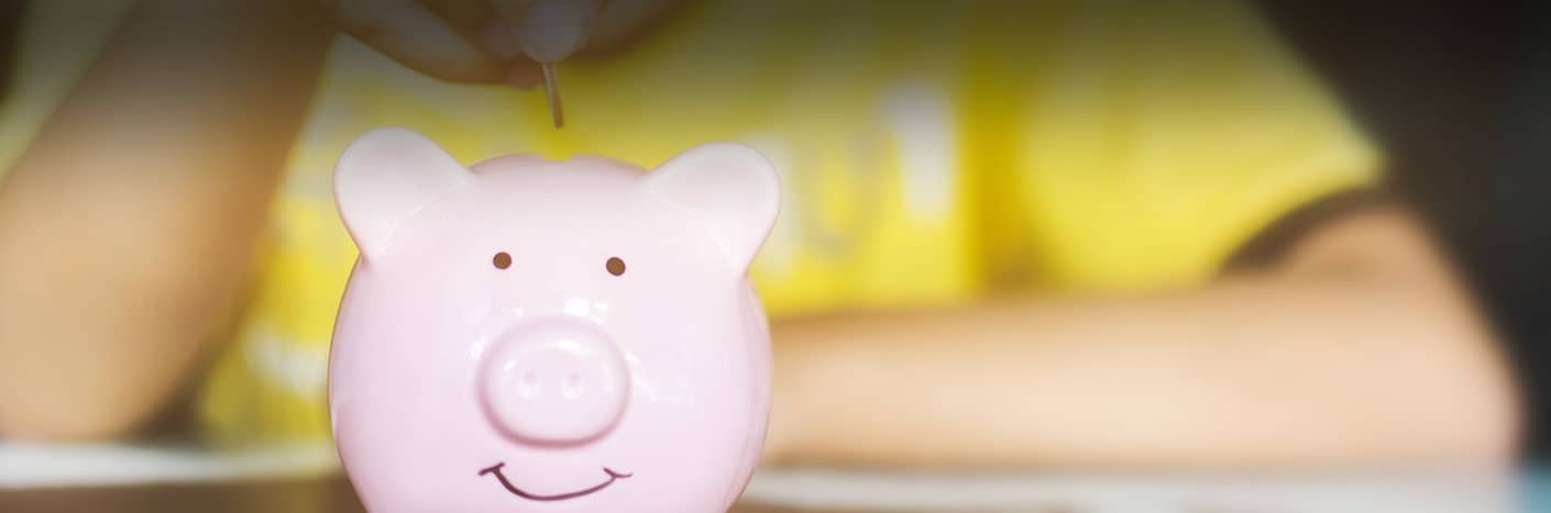 Eine Person legt eine Münze in ein lächelndes rosa Sparschwein auf einem Tisch.