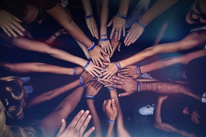 Eine Gruppe von Menschen reicht sich in der Mitte die Hände und trägt passende blaue Armbänder, die Einigkeit und Teamwork symbolisieren.