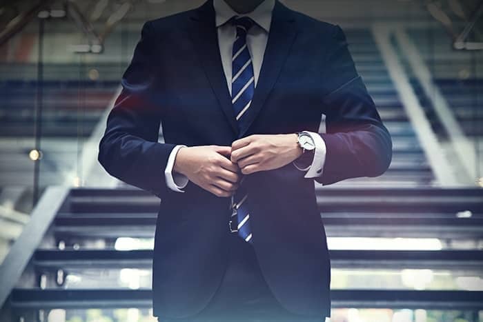 Eine Person in Anzug und Krawatte knöpft ihre Jacke zu und steht vor einer Treppe in einem modernen Gebäude.