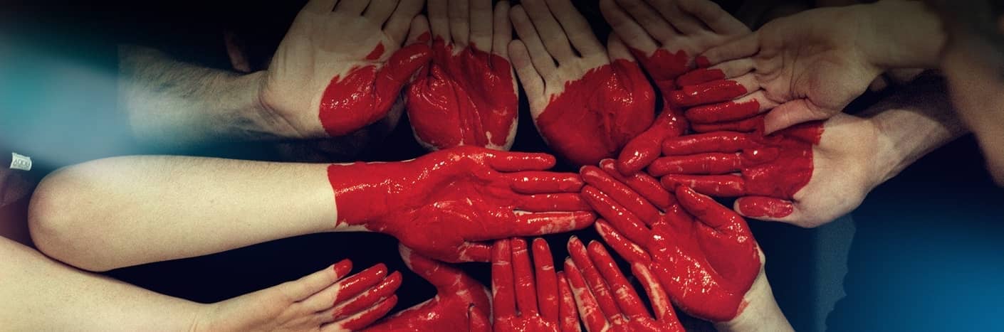 Mehrere rot bemalte Hände sind vor einem dunklen Hintergrund zu einem Herz zusammengefügt.