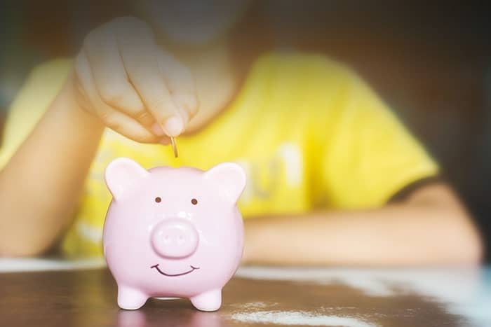 Eine Person in einem gelben Hemd wirft eine Münze in ein rosa Sparschwein auf einem Tisch.