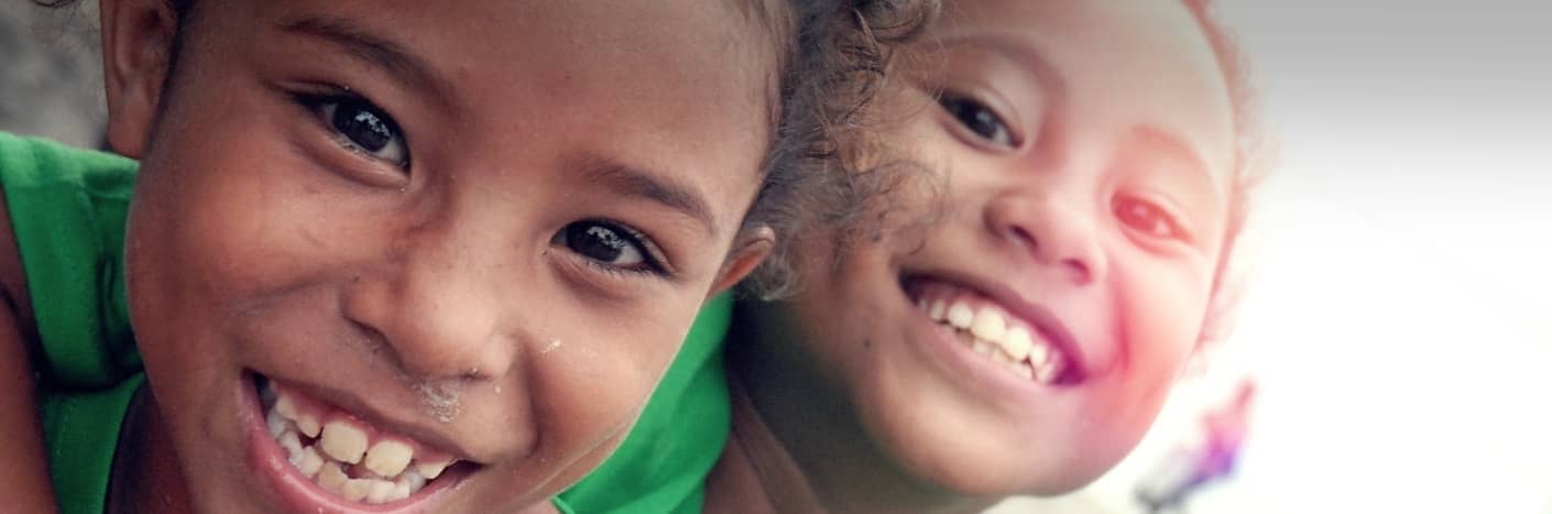 Zwei Kinder in grünen Hemden, die eng beieinander stehen und fröhlich lächeln, vor einem weichen, hellen Hintergrund.