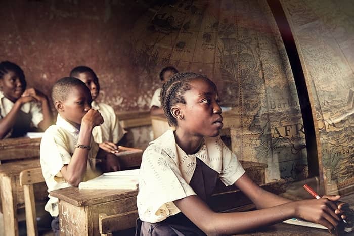 Uniformierte Schüler sitzen an hölzernen Schreibtischen in einem Klassenzimmer und hören aufmerksam zu, während eine alte Karte von Afrika eingeblendet wird.