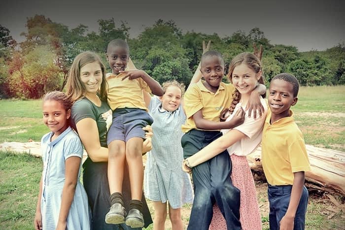 Sechs lächelnde Kinder und zwei Frauen posieren gemeinsam im Freien auf einer Wiese, mit Bäumen und umgestürzten Baumstämmen im Hintergrund.