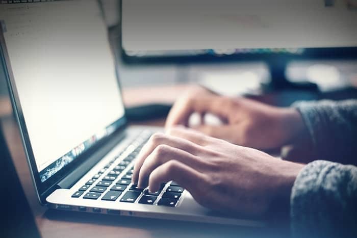 Hände, die auf einer Laptoptastatur tippen, mit verschwommenen Bildschirmen im Hintergrund.