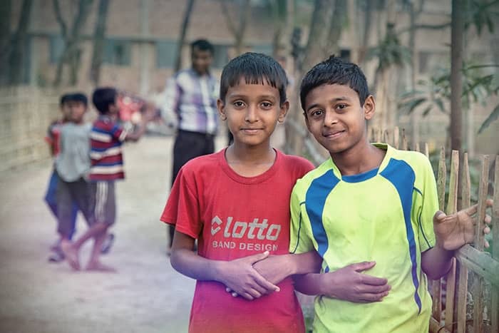 Zwei Jungen stehen dicht beieinander und lächeln in die Kamera, während im Hintergrund andere Kinder im Freien spielen.