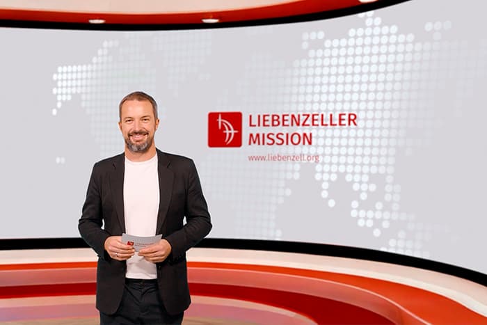 Ein lächelnder Mann im Anzug steht in einem modernen Studio mit dem Logo der Liebenzeller Mission auf einem Bildschirm hinter ihm.