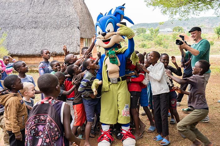 Kinder versammeln sich aufgeregt um eine Person in einem Sonic the Hedgehog-Kostüm im Freien und umarmen sie, während ein Mann Fotos macht.