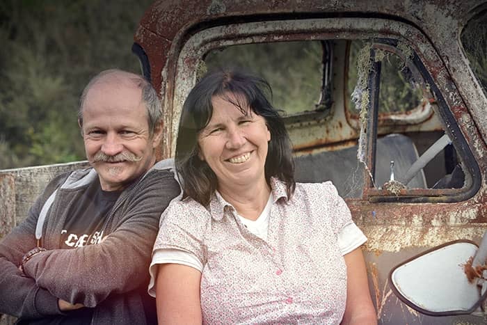 Ein lächelnder Mann und eine lächelnde Frau sitzen gemeinsam vor einem alten, verrosteten Auto im Freien.