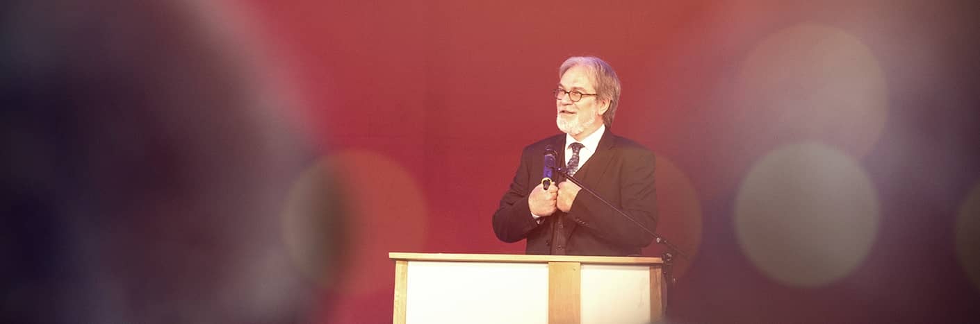 Ein älterer Mann im Anzug spricht an einem Podium mit einem Mikrofon vor einem verschwommenen, warm beleuchteten Hintergrund.