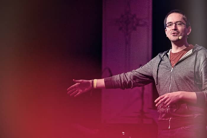 Ein Mann mit Headset und grauem Kapuzenpulli gestikuliert mit einem ausgestreckten Arm, während er auf einer Bühne mit lila Beleuchtung spricht.