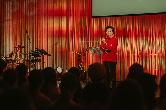 Ein Redner in einem roten Hemd wendet sich an das Publikum auf der Bühne, während im Hintergrund orangefarbene Beleuchtung und Musikinstrumente zu sehen sind.