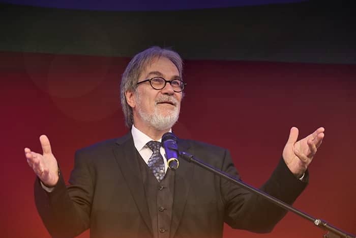 Ein Mann im Anzug und mit Brille spricht an einem Mikrofon und gestikuliert mit offenen Armen vor einem roten Hintergrund.