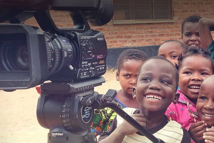 Eine Gruppe lächelnder Kinder steht dicht beieinander hinter einer Videokamera im Freien in der Nähe eines Backsteingebäudes.