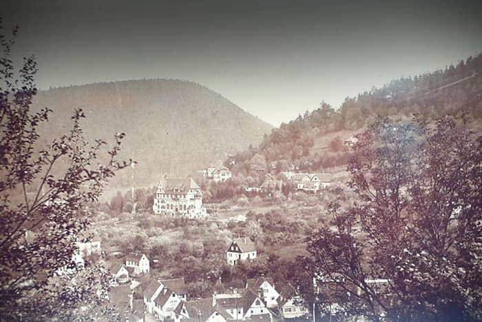 Sepiafarbenes Foto eines Bergdorfs mit verstreuten Häusern und Bäumen vor der Kulisse bewaldeter Berge.