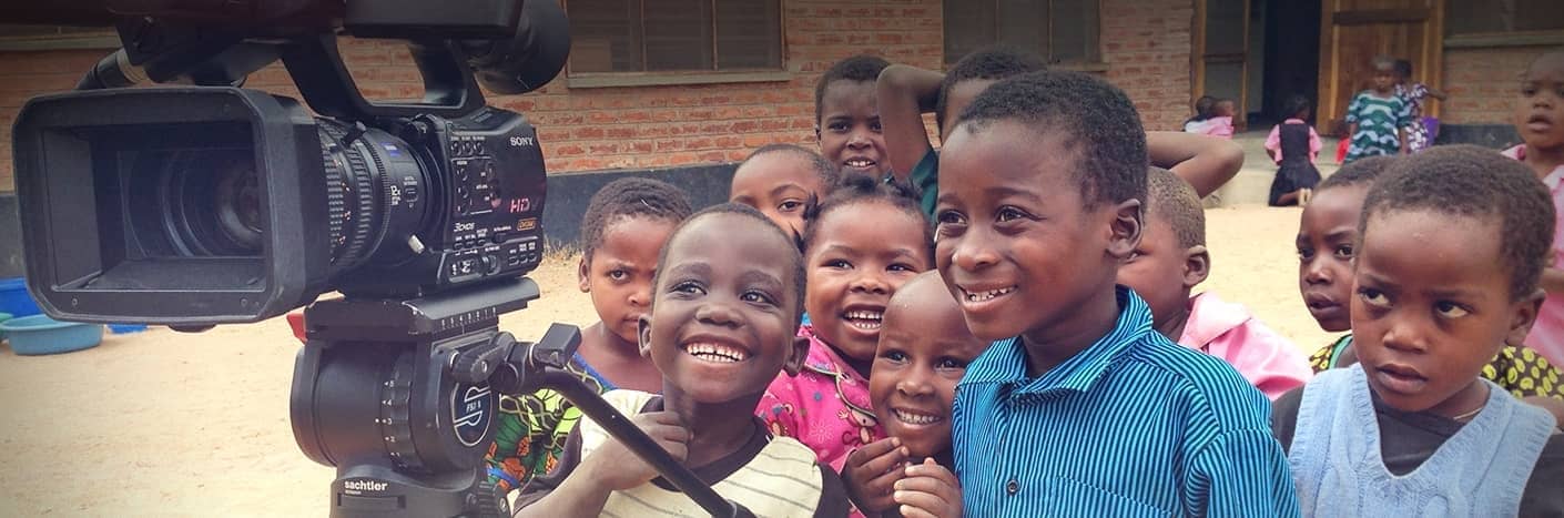 Eine Gruppe lächelnder Kinder versammelt sich im Freien um eine Videokamera und zeigt sich aufgeregt und neugierig.