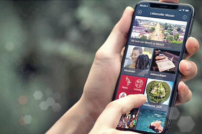 Eine Person hält ein Smartphone, auf dem eine missionsbezogene App mit verschiedenen Bildern und Menüoptionen angezeigt wird.