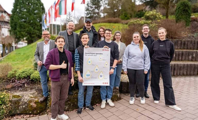 Eine Gruppe von elf Personen steht im Freien, lächelt und hält ein Plakat mit Texten und Diagrammen über HIV und Blutspenden.