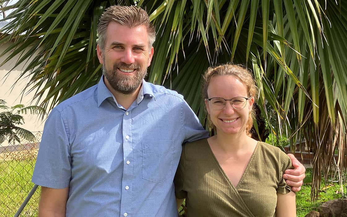Ein Mann und eine Frau lächeln im Freien und stehen zusammen vor einer großen Palme.