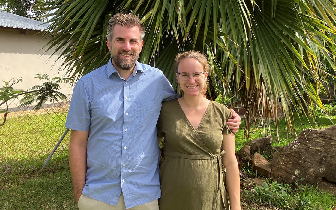 Ein Mann und eine Frau lächeln im Freien und stehen zusammen vor einer großen Palme und grünem Laub.