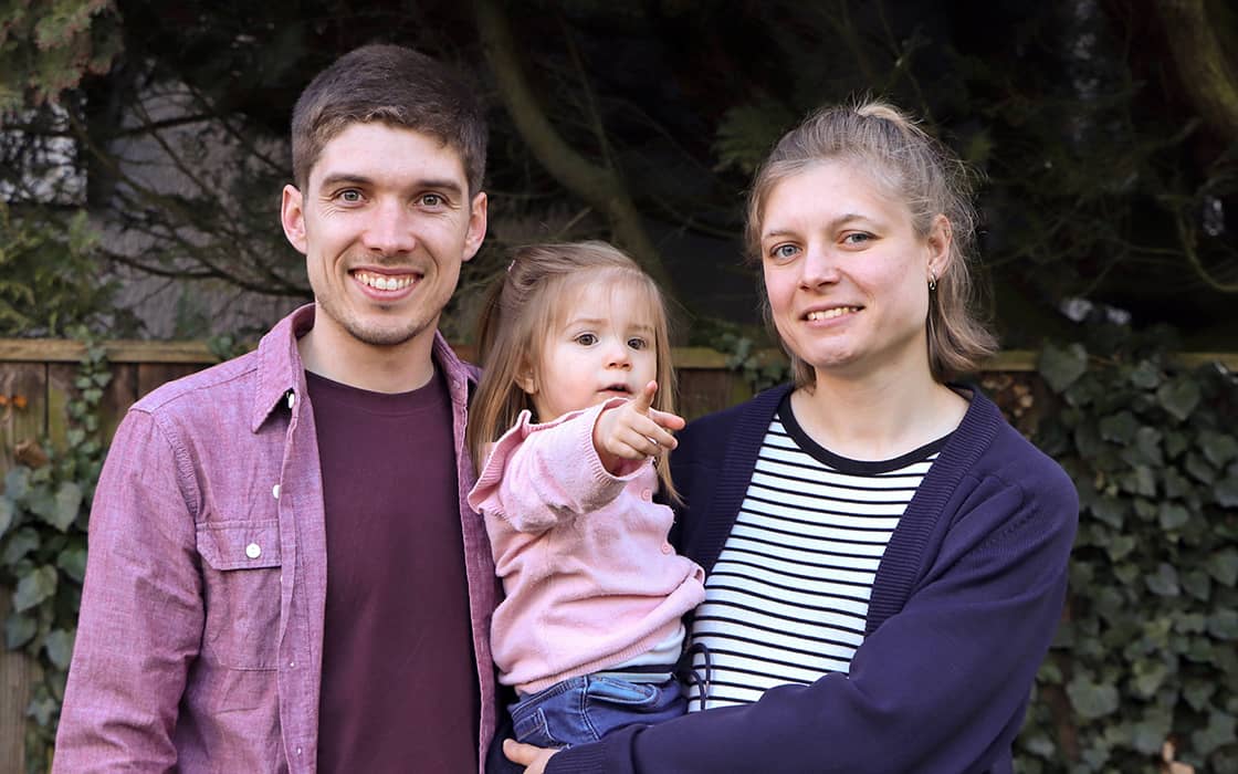 Ein Mann und eine Frau lächeln, während sie im Freien mit einem jungen Mädchen stehen, das in die Kamera zeigt.