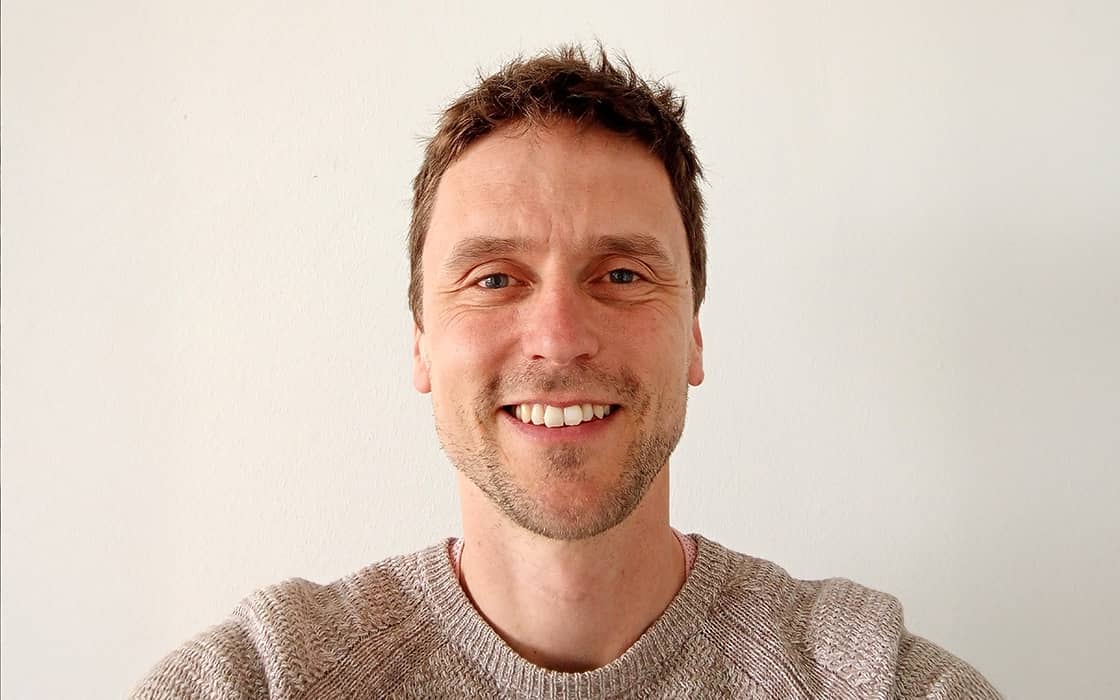 Ein Mann mit kurzen braunen Haaren lächelt in die Kamera und trägt einen hellgrauen Pullover vor einem schlichten weißen Hintergrund.