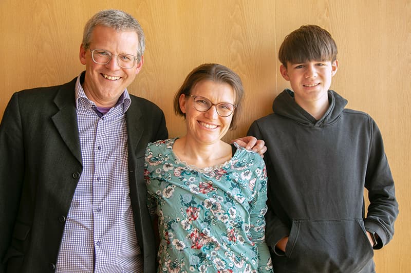 Drei Personen, zwei Erwachsene und ein Jugendlicher, stehen lächelnd zusammen vor einer Holzwand.