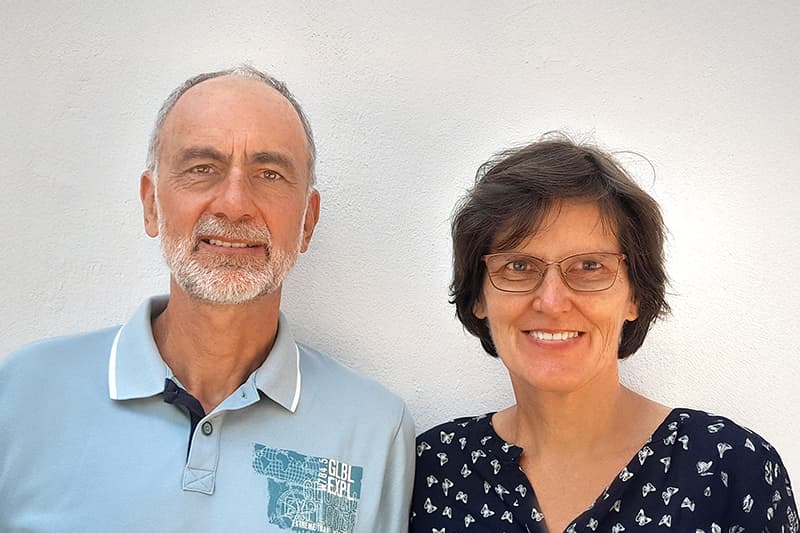 Ein lächelnder älterer Mann in einem hellblauen Polohemd und eine Frau mit Brille stehen gemeinsam vor einer schlichten weißen Wand.