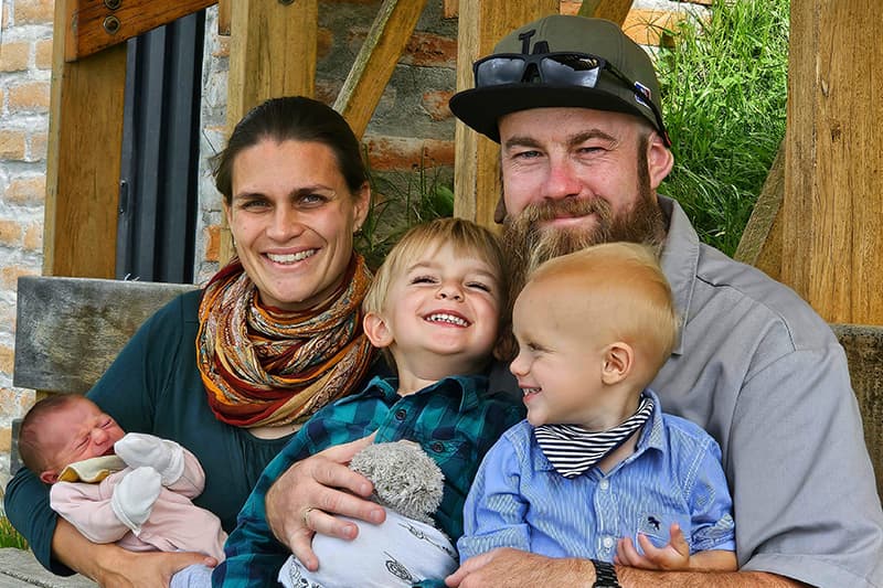 Lächelnde fünfköpfige Familie posiert im Freien; die Eltern halten drei kleine Kinder, darunter ein in eine Decke eingewickeltes Baby.