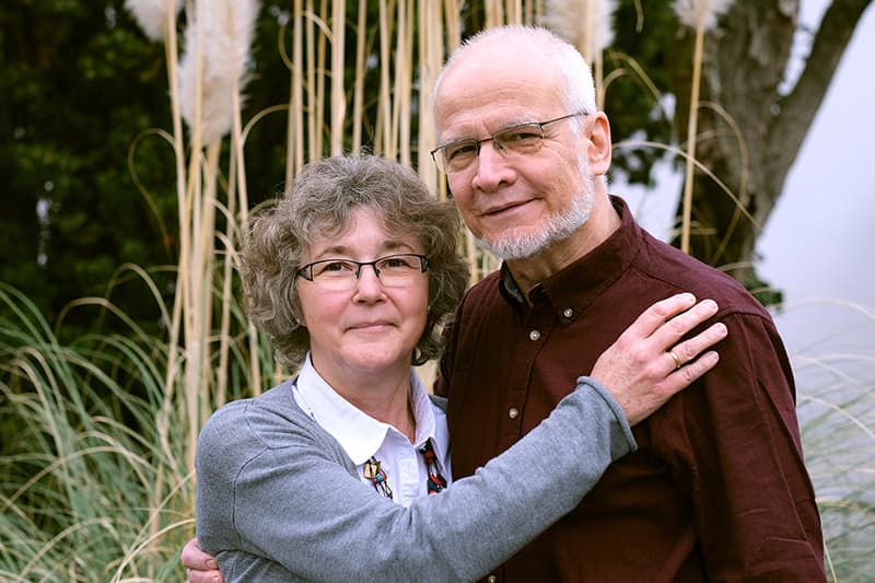 Ein älteres Paar steht im Freien dicht beieinander, lächelnd, mit hohen Gräsern und Bäumen im Hintergrund.