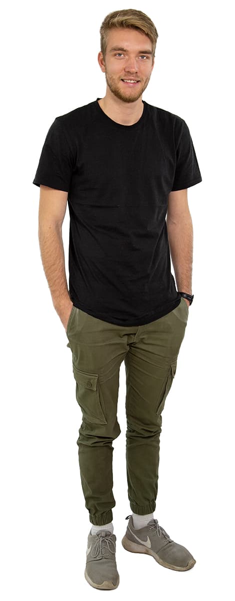 Stehender Mann, schwarzes T-Shirt, olivgrüne Cargohose, graue Turnschuhe, Hände in den Taschen, lächelnd.