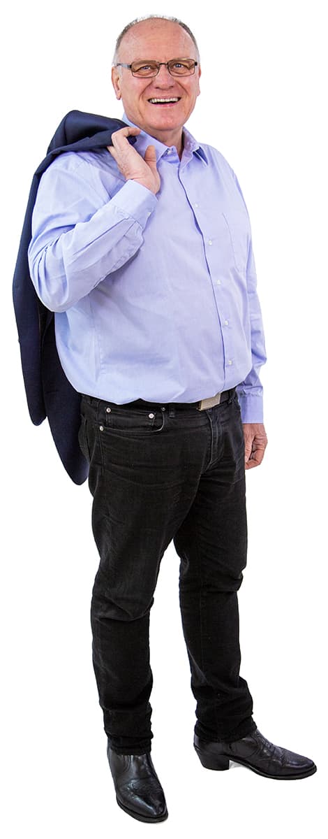 Lächelnder Mann in hellblauem Hemd, schwarzer Hose und Stiefeln, der eine Jacke über der Schulter trägt, steht auf weißem Hintergrund.