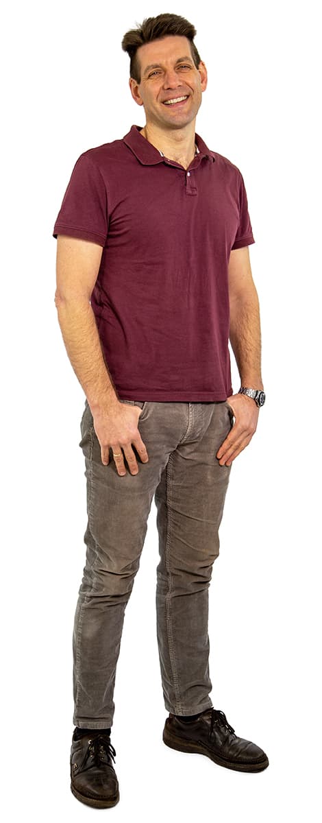 Stehender, lächelnder Mann mit kastanienbraunem Poloshirt, grauen Jeans, schwarzen Schuhen und einer Uhr auf weißem Hintergrund.