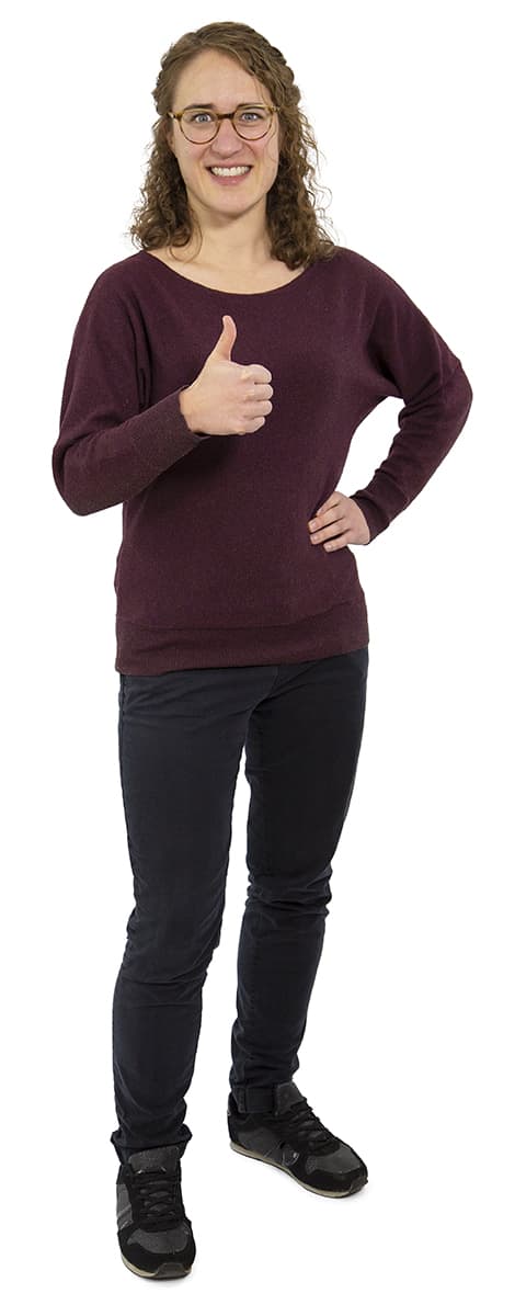 Frau mit lockigem Haar, die eine Brille, einen kastanienbraunen Pullover und eine schwarze Hose trägt, lächelt und zeigt den Daumen hoch.