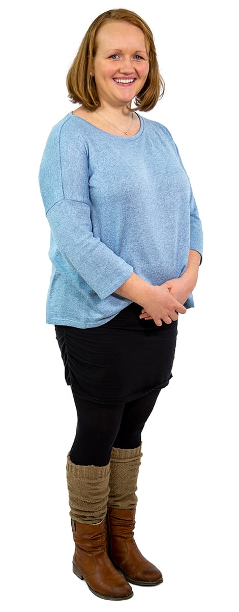 Lächelnde Frau, die einen hellblauen Pullover, einen schwarzen Rock, schwarze Leggings und braune Stiefel trägt und vor einem weißen Hintergrund steht.