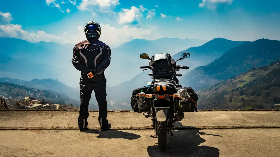 Ein Motorradfahrer steht neben seiner Maschine und blickt auf eine malerische Berglandschaft unter blauem Himmel.