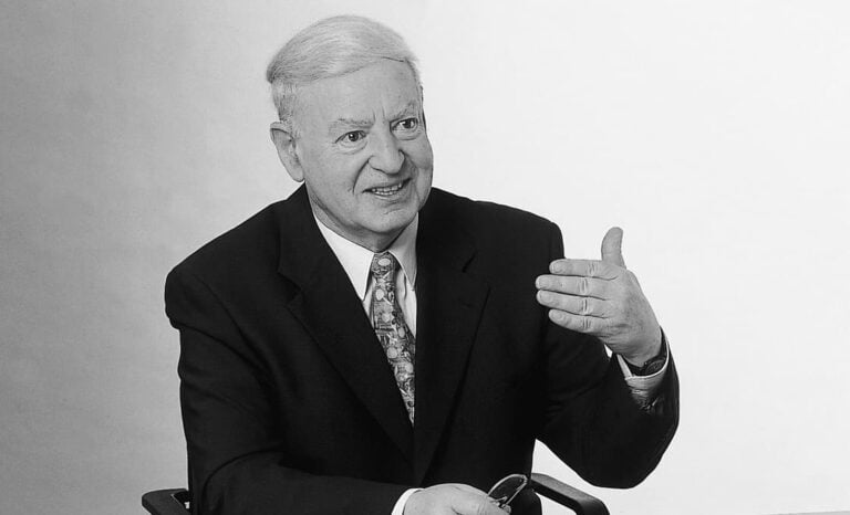 Älterer Mann im Anzug sitzt und gestikuliert mit seiner rechten Hand, in der linken Hand hält er eine Brille, vor einem schlichten Hintergrund.