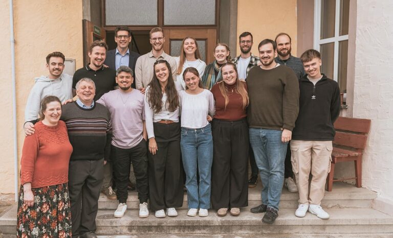 Fünfzehn lächelnde Menschen stehen auf einer Freitreppe vor einem Gebäude für ein Gruppenfoto zusammen.
