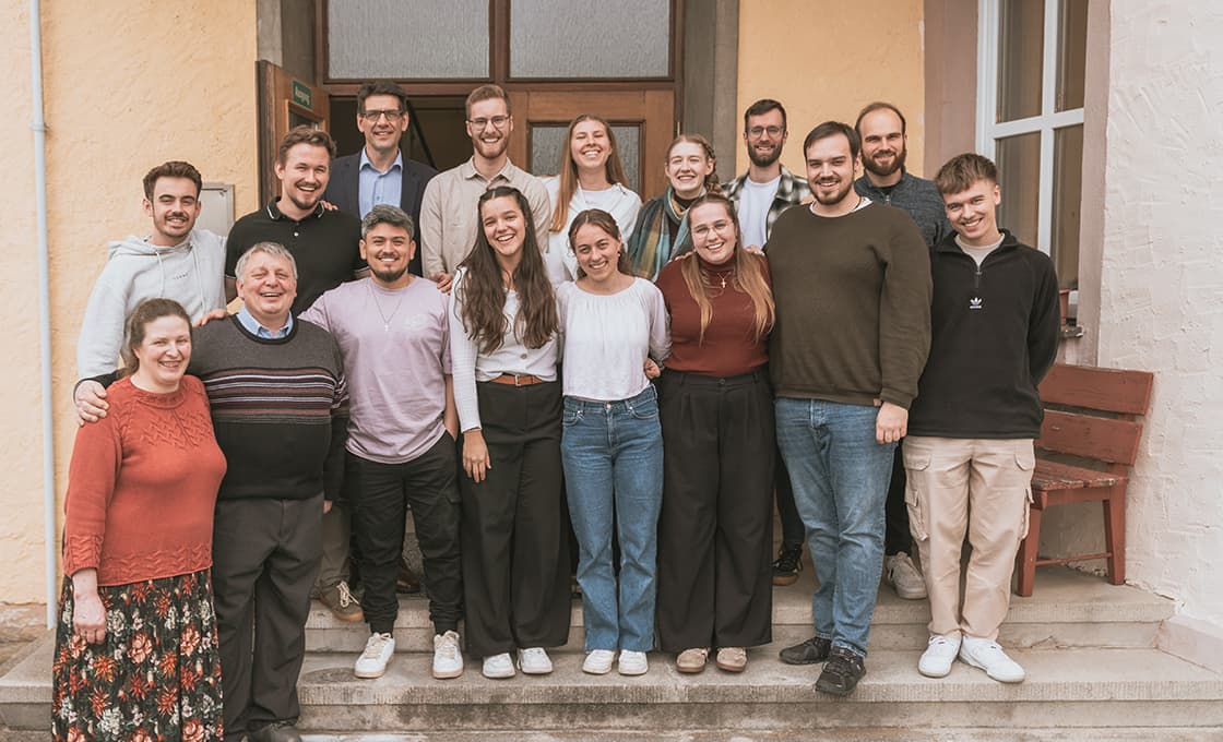 Fünfzehn lächelnde Menschen stehen auf einer Freitreppe vor einem Gebäude für ein Gruppenfoto zusammen.