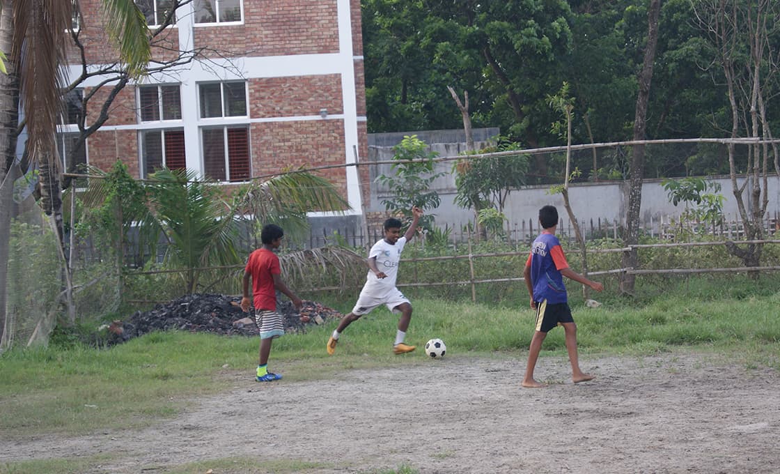 Drei Jungen spielen Fußball auf einer Wiese in der Nähe eines Gebäudes, mit Bäumen und einem Zaun im Hintergrund.
