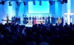Ein Chor steht in einer Kirche auf der Bühne, dem Publikum zugewandt, mit blauer Beleuchtung und einem großen beleuchteten Kreuz.