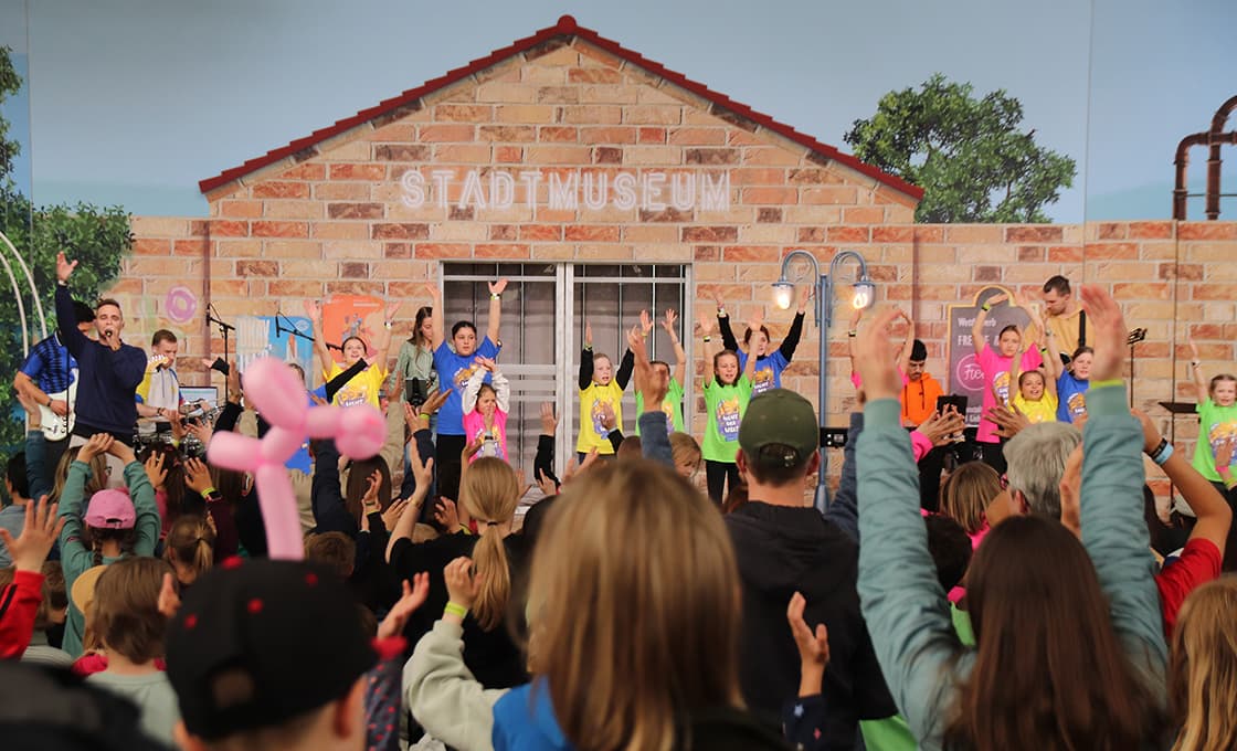 Eine Gruppe von Kindern spielt energisch auf der Bühne, während eine Menschenmenge vor einem Gebäude mit der Aufschrift "Stadtmuseum" zusieht.
