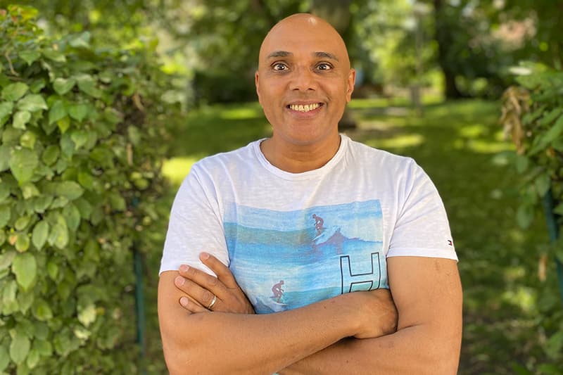Ein lächelnder Mann mit rasiertem Kopf steht mit verschränkten Armen im Freien und trägt ein weißes T-Shirt mit einem Surfer-Aufdruck.
